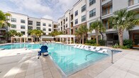 This outdoor amenity space is centered around a generously sized swimming pool with crystal-clear blue water, framed by a well-maintained travertine tile deck that provides ample room for relaxation and recreation. Rows of white lounge chairs line the poolside, offering residents comfortable spaces to unwind, while a classic white pergola at the far end of the pool provides a shaded gathering area. A dedicated lounge zone adjacent to the pool deck features additional sling-style chaise lounges alongside a private cabana with draped curtains, creating an intimate and resort-like atmosphere. Tall palm trees are thoughtfully integrated throughout the space, enhancing the tropical ambiance and providing natural shade. The pool area is enclosed by wrought iron fencing and bordered by manicured landscaping with tropical plantings and decorative rock mulch. Accessibility is a priority, with a pool lift available for ease of entry. Clearly posted pool policies ensure a safe and organized environment for all users. Covered walkways and secured entry points connect the pool deck to the surrounding multi-story building, which features large windows, private balconies, and a contemporary facade blending stucco and brick. The overall design evokes a relaxed, upscale outdoor living experience.