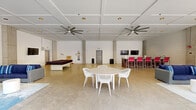 Located at 201 Jefferson St N in Huntsville, Alabama, this single-floor amenity space is designed to impress with a blend of modern industrial and coastal-inspired design elements. The centerpiece is a spacious, open-plan activity hall featuring polished concrete floors, soaring coffered ceilings, and oversized ceiling fans that lend both form and function to the airy environment. Exposed concrete columns frame the room, while recessed lighting and a wall-mounted flat-screen TV add contemporary convenience. A bar-height counter with vibrant red seating provides a social gathering spot, and a large padded platform offers additional flexible use of the space.

Adjacent to the main hall is a comfortable lounge area decorated with navy blue sofas and wicker chairs accented by bold pink and striped throw pillows. Shiplap-style white walls and a decorative guitar wall piece contribute to a relaxed, creative atmosphere. Brick accent columns add a touch of warmth and character throughout.

The outdoor area extends the experience with a beautifully appointed resort-style pool, framed by neatly arranged turquoise lounge chairs, lush tropical plantings, and tall cypress trees. A pergola structure and thoughtfully landscaped surroundings create a serene retreat, all set against the backdrop of a striking modern multi-story building.