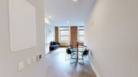 Located at 210 Main St, Unit 610 in Hackensack, NJ, this one-bedroom, one-bathroom residence blends industrial character with modern finishes across a well-organized single-floor layout. The living area is a standout, showcasing original exposed brick walls paired with large double-hung windows that invite natural light and frame views of the surrounding streetscape. Light-toned hardwood flooring runs throughout, unifying the space with warmth and continuity. The kitchen is sleek and fully equipped, featuring glossy white cabinetry that extends to the ceiling, a subway tile backsplash, stainless steel appliances including a range, microwave, and dishwasher, and a dedicated dining area nearby. The bedroom continues the exposed brick aesthetic, with two windows providing cross-ventilation and natural light. A dedicated walk-in closet serves the bedroom, complete with wooden hanging rods and built-in shelving for generous storage. The bathroom is cleanly finished with white subway tile surrounding a full soaking tub and shower combo, accented by a decorative mosaic tile border, a wood-toned vanity, and a large mirror. Recessed lighting and neutral wall tones throughout contribute to a bright, contemporary ambiance.