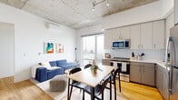 Located in Chicago's Lincoln Park neighborhood, this contemporary unit at 1450 N Dayton St showcases a thoughtfully designed open-concept layout that seamlessly connects the living, dining, and kitchen areas. The space is defined by striking exposed concrete ceilings with integrated track lighting, warm light wood flooring throughout, and crisp white walls that create a clean, modern industrial aesthetic. Large windows flood the living area with natural light while offering sweeping city views, enhancing the airy atmosphere. A wall-mounted split AC unit ensures year-round comfort.

The kitchen is well-appointed with two-tone gray cabinetry, classic white subway tile backsplash, and a full suite of stainless steel appliances including a refrigerator, range, microwave, and dishwasher. The open dining area accommodates a full dining table, making it ideal for entertaining.

Multiple bedrooms provide private retreats, each featuring light wood flooring, large windows with city views, and exposed concrete columns that echo the industrial design language of the main living space. The bathrooms are finished with white subway tile, a glass-enclosed shower, gray cabinetry with brushed gold hardware, and warm wood flooring, delivering a cohesive and polished look throughout.