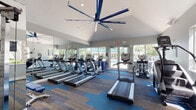 This fitness center is thoughtfully designed to support a wide range of workout routines in a bright, welcoming environment. The main cardio area features a row of treadmills and elliptical machines positioned beneath a striking large-blade ceiling fan, with floor-to-ceiling mirrors amplifying the sense of space. Glass doors open directly to an outdoor pool area, flooding the room with natural light and providing refreshing views of the surrounding landscape, including lush palm trees.

The vaulted ceiling adds an open, airy quality to the main workout floor, which is finished with a modern patterned carpet in neutral and blue tones. A dedicated strength training section houses a multi-station cable machine, a leg press, and additional resistance equipment, all positioned beneath large windows that bring in ample daylight and outdoor views.

A separate functional training area features hardwood-style flooring and includes stability balls in multiple sizes, a full set of medicine balls, a rack of dumbbells, a BOSU balance trainer, and a wall-mounted display screen for on-demand workout classes. Personal storage lockers are conveniently available within the facility. Together, these zones create a comprehensive, well-organized fitness space suited for both cardio conditioning and full-body strength training.