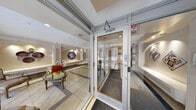 This single-floor space functions as a building lobby and common area, thoughtfully designed with classical elegance and attention to architectural detail. The entrance is framed by full-height glass panels and an automatic door system, flooding the interior with natural light and offering clear sightlines to the surrounding streetscape.

Immediately past the entrance, a bright lounge area features large floor-to-ceiling windows on multiple sides, providing panoramic outdoor views. This sun-filled sitting area is finished with light ceramic tile flooring and serves as a welcoming reception point, furnished with classic upholstered chairs, a glass-topped table, and decorative indoor plants.

The hallway transitions seamlessly from tile to carpet, guided by structured white pilasters and detailed crown molding that run throughout. Arched wall niches are illuminated with accent lighting and display curated decorative artwork, adding a gallery-like character to the corridor. A prominent circular wall piece is spotlit within one of the arches, serving as a visual focal point.

The space continues into a secondary hallway leading to an elevator with polished metal doors. Built-in benches and subtle recessed lighting contribute to a composed, refined ambiance throughout. The overall design conveys a professional and polished aesthetic with consistent warm neutral tones.