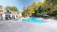 Located in Tucker, Georgia, this outdoor pool area offers a well-appointed recreational retreat designed for relaxation and leisure. The centerpiece is a generously sized swimming pool with clear blue water, framed by an expansive stamped concrete deck that provides ample space for sunbathing and socializing. Surrounding the pool, lounge chairs and seating arrangements are thoughtfully positioned to take advantage of the natural light and serene wooded backdrop of mature trees that provide shade and privacy.

A charming pergola with seating adds a shaded gathering spot, perfect for outdoor dining or relaxing out of the sun. Multiple patio umbrellas are stationed around the deck, offering additional shade options for guests. The pool area is enclosed by a black wrought iron fence, ensuring both security and an open, airy aesthetic.

Adjacent to the pool, a poolside building features a classic white shiplap exterior with large multi-pane windows and stone masonry accents, housing restroom facilities and a water fountain for convenience. The building's architectural design complements the residential character of the community. Altogether, this outdoor space combines functional amenities with natural beauty, creating an inviting environment for recreation and community gathering throughout the warmer months.