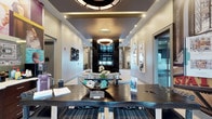 This elegantly designed single-floor communal space presents a cohesive blend of contemporary style and functional comfort across several distinct zones. The main lounge area serves as the centerpiece, anchored by a dramatic floor-to-ceiling dark tile fireplace surround with a wall-mounted television above. Generous seating arrangements—including plush sofas, upholstered armchairs, and accent ottomans—are arranged atop boldly patterned area rugs featuring wave-like designs in deep brown and white tones.

Overhead, large circular pendant fixtures with warm, layered lighting illuminate the space, complemented by recessed lighting embedded in tray and coffered ceiling details. Light-toned large-format tile flooring extends throughout, creating a clean, open feel. A dedicated café-style dining nook features bistro tables with sculptural metallic pedestal bases and leather-upholstered bar stools, positioned near large windows that allow abundant natural light to filter in through woven roman shades.

The entry area showcases decorative wall art including circular metallic disc sculptures and abstract paintings, reinforcing the space's curated aesthetic. A game area with a billiard table is visible in the background, adding a recreational dimension. The overall ambiance is polished and upscale, designed to encourage both social interaction and relaxed lounging.