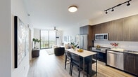 Located in Miami, FL, this well-appointed one-bedroom, one-bathroom residence offers a thoughtfully designed open-concept layout across a single floor. Light-toned hardwood-style flooring flows throughout, creating a cohesive and airy feel.

The combined kitchen and living area serves as the heart of the home. The kitchen is outfitted with dark wood cabinetry, stainless steel appliances including a French door refrigerator and range, a tile backsplash, and a central island with a quartz countertop and seating — ideal for casual dining. The living area is bright and spacious, anchored by floor-to-ceiling sliding glass doors that open directly to a private balcony with expansive views of the Miami skyline and surrounding greenery. A ceiling fan adds comfort overhead.

The bedroom is a serene retreat, featuring large picture windows with panoramic outdoor views, a ceiling fan, and direct access to a dedicated walk-in closet with built-in dark wood shelving and hanging storage.

The bathroom is finished in a clean, modern aesthetic with large-format white tiles, a frameless glass walk-in shower with a rain showerhead, matte black fixtures, and a quartz vanity surface.

A hallway connects the living spaces efficiently, completing this polished and functional Miami residence.