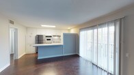 This single-floor space offers a thoughtfully designed open-plan layout that seamlessly connects the kitchen and living areas, creating a cohesive and functional environment. The kitchen is well-appointed with crisp white cabinetry that extends to the ceiling, providing ample storage. Stainless steel appliances, including a refrigerator and built-in oven, lend a modern and practical character to the space. A central island with a light blue base serves as a natural dividing feature between the kitchen and the adjoining living area, offering both workspace and a visual focal point. The rich, dark hardwood flooring grounds the space and adds warmth that contrasts beautifully with the light cabinetry and walls. The living area is bathed in natural light, courtesy of expansive floor-to-ceiling sliding glass doors dressed with sheer vertical blinds that filter soft, diffused daylight throughout the room. The neutral wall tones and clean recessed and panel lighting overhead contribute to a bright, airy ambiance. Overall, the space strikes a balance between practicality and a welcoming, open atmosphere suited for comfortable everyday living.