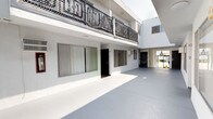 This multi-unit building presents a striking white stucco exterior that reflects the abundant natural sunlight characteristic of its warm climate setting. The structure is arranged around an open central courtyard, which serves as the primary common area and circulation space. The courtyard is finished with a smooth, light-colored concrete surface that amplifies the brightness of the space and creates a clean, airy atmosphere.

Ground-floor units line the courtyard, each accessed via individual dark-framed entry doors and accompanied by large sliding windows fitted with vertical blinds, allowing residents to control light and privacy. Wall-mounted HVAC units are visible between windows, providing climate control for each unit.

Above, an upper level features a prominent balcony wrapped in ornate, geometric wrought iron railings — a decorative architectural detail that adds character and visual interest to the otherwise minimalist facade. The upper-level balcony overlooks the courtyard below, contributing to a sense of vertical openness.

At the building's perimeter, decorative white masonry balustrades with an open latticework design frame the exterior walkways and staircase. A glimpse of a mature palm tree beyond the property boundary hints at a classic Southern California setting. Fire safety equipment is thoughtfully integrated into the courtyard walls, reflecting attention to building standards throughout the property.
