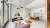 Located in the heart of Durham, NC, this elegantly designed lobby at 321 W Corporation Street offers a refined first impression across a thoughtfully arranged single-floor layout. The living room lounge area is an immediate showstopper, anchored by dramatic floor-to-ceiling windows that flood the space with natural light and frame vibrant street views of the surrounding urban neighborhood. A striking geometric chandelier hangs overhead, complementing the contemporary seating arrangement of plush sofas, accent chairs, and organically shaped wooden coffee tables atop a neutral area rug. The overall palette is crisp and modern, with white walls and polished concrete flooring enhancing the airy, open feel. Transitioning through the lobby, four distinct hallways feature textured concrete-finish walls in a warm gray tone, illuminated by recessed and track lighting that highlights a rotating gallery of large-format framed artwork. Light hardwood flooring and sleek black benches lend a gallery-like sophistication to these corridors. The dining room offers an intimate, functional space with soaring ceilings, a warm walnut table surrounded by modern upholstered chairs, and a wall-mounted shelving system displaying curated objects and books. Framed artwork adds a finishing touch, making this room ideal for meetings or private gatherings. Together, these spaces create a lobby that is both welcoming and architecturally refined.