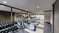 This two-floor fitness facility is thoughtfully designed to accommodate a wide range of exercise needs. The gym features a robust selection of cardio equipment, including multiple treadmills and elliptical trainers arranged in a spacious, organized layout that allows for comfortable movement between machines. A dedicated free weights area includes a full rack of dumbbells, catering to strength training routines of varying intensity. Large mirrored panels line the walls, visually expanding the space and providing exercisers with proper form feedback during workouts. Recessed lighting throughout the ceiling ensures the space is consistently well-lit, creating an energizing atmosphere. The flooring combines durable, cushioned surfaces with striped detailing, offering both safety and aesthetic appeal. Natural light filters in through frosted windows, adding a bright, airy quality to the environment. The clean, neutral color palette — with light walls, dark trim accents, and grey equipment — contributes to a focused and professional ambiance. Overall, the facility is designed with functionality and comfort in mind, making it a practical and motivating space for daily fitness activities.