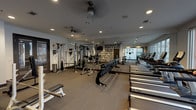 This fitness center is organized across a single floor and divided into distinct functional zones designed to support a comprehensive workout experience. The main exercise area is generously sized, featuring a full lineup of TRUE-brand treadmills and cardio machines arranged in a row along one side, offering views through large street-facing windows that fill the space with natural light. Opposite the cardio equipment, a dedicated free weights section includes a multi-tiered dumbbell rack, adjustable benches, and a cable resistance machine, providing ample options for strength training. A large floor-to-ceiling mirror runs along one wall, visually expanding the space, while a bold motivational mural — featuring phrases such as "Be Fearless," "Have a Vision," and "Believe" — energizes the environment. Ceiling fans and recessed lighting maintain a comfortable atmosphere throughout. A separate studio room, accessible through sleek frosted-glass sliding doors, features soaring double-height ceilings, mirrored walls, and its own sound system, making it well-suited for group fitness or cycling classes. The flooring transitions between durable rubber and carpet tile, reinforcing the functional yet polished character of the overall space.