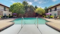 This outdoor amenity space in Suwanee, Georgia offers a well-appointed recreational environment centered around a generously sized swimming pool with clean tile surroundings and stainless steel entry rails. The pool is framed by mature trees that provide natural shade and a serene backdrop, while colorful umbrella-shaded seating areas invite relaxation along the pool deck.

Adjacent to the pool, a spacious open-air courtyard serves as a central gathering area, featuring large decorative planters, umbrella-shaded tables, and ample concrete patio space ideal for outdoor socializing. The courtyard is flanked by a multi-story building with brick and stone facade accents, individual balconies, and well-maintained landscaping that adds warmth to the overall setting.

A covered pavilion area provides a shaded retreat supported by classic brick columns and equipped with ceiling fans for year-round comfort. This versatile space includes seating, a small table, and decorative wall accents, making it suitable for both casual gatherings and quiet relaxation. The pavilion connects seamlessly to the surrounding outdoor areas, creating a cohesive and functional amenity complex. Together, these spaces deliver a welcoming, resort-inspired outdoor experience beneath the open Georgia sky.