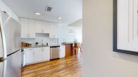 This single-floor urban residence showcases a thoughtfully designed open layout with warm hardwood flooring and a clean, contemporary aesthetic throughout. The kitchen is a standout feature, equipped with stainless steel appliances including a refrigerator, range, built-in microwave, and dishwasher. Sleek white cabinetry extends to the ceiling, complemented by a subway tile backsplash and a peninsula with a dark countertop and bar seating beneath a modern pendant light — ideal for casual dining.

The living area benefits from floor-to-ceiling black-framed glass doors that open directly onto a private balcony, flooding the space with natural light and offering sweeping city skyline views. Exposed concrete structural columns add an industrial character that contrasts beautifully with the bright white walls.

The bedroom features tall windows with additional city views, a ceiling fan, and a walk-in closet with built-in wire shelving. Two bathrooms serve the unit: one with a soaking tub and shower combination with a white tile surround and a dual-sink vanity, and a second with a shower and a streamlined white vanity. The balcony overlooks a pool courtyard, adding to the elevated urban living experience.