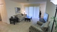 This single-floor space centers around a well-appointed living room designed for both comfort and functionality. The room features light neutral-toned walls complemented by plush wall-to-wall carpeting, creating a warm and inviting atmosphere throughout. A large sliding glass door dressed with vertical blinds serves as the focal point of the space, allowing abundant natural light to filter in while providing access to the outdoors. The living area is generously sized, comfortably accommodating a full sofa with decorative accent pillows, a tufted ottoman that doubles as a coffee table, and additional seating in the form of a patterned accent chair. A dark wood side table and matching console table add contrast against the lighter elements of the room. Subtle décor, including a landscape-inspired wall canvas and a small table lamp, contribute to the space's understated, residential character. A glimpse into an adjoining area is visible beyond the living room, suggesting a connected layout that flows naturally between spaces. Overall, the space presents a clean, move-in-ready environment with a calm and approachable aesthetic.