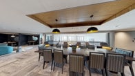 This thoughtfully designed single-floor amenity space offers a variety of distinct zones that cater to both communal gatherings and quiet relaxation. The centerpiece of the space is a generous open-plan lounge and dining area featuring a large dark wood communal table surrounded by upholstered striped chairs, all anchored beneath a striking wood-paneled ceiling detail with bold black-and-gold dome pendant lights. The herringbone-patterned hardwood-style flooring flows throughout, lending warmth and continuity across the space. Adjacent to the dining area, an open lounge zone provides an assortment of contemporary swivel chairs with yellow upholstered accents, creating vibrant pops of color against the neutral palette. Large windows along the perimeter flood the space with natural light and offer views of the surrounding neighborhood. A dedicated media lounge features deep sectional seating, textured stone accent walls, and a large wall-mounted flat-screen television set above dark cabinetry with a light quartz countertop. Artistic touches are woven throughout, including abstract canvas artwork and sculptural wood mosaic wall panels. A more intimate lounge nook with dark walls, a bookshelf, and relaxed seating rounds out the space, providing a cozy retreat. The overall ambiance is modern and polished, blending warm materials with contemporary design elements.