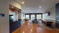 Located at 15155 Richmond Ave in Houston, Texas, this single-floor community clubhouse offers a well-appointed gathering space designed for both recreation and productivity. The main area showcases striking polished concrete floors that flow throughout, complemented by recessed lighting and a warm neutral color palette accented by deep navy blue walls and wood trim details.

The expansive game and lounge area serves as the centerpiece of the space, featuring a full-size pool table, a shuffleboard table, and a casual dining table with bench seating. A wall-mounted flat-screen television adds to the entertainment appeal, while large sliding glass doors and generously sized windows flood the room with natural light and offer views of the surrounding outdoor areas and landscaping.

The kitchenette is equipped with warm wood cabinetry, a slate tile backsplash, dark granite countertops, a black refrigerator, a microwave, and a peninsula with a navy blue base — ideal for hosting gatherings and events. Adjacent to the main lounge, a dedicated business center provides a quiet workspace complete with a computer workstation and carpeted flooring framed by classic wood-trimmed glass doors. The overall ambiance is welcoming and versatile, making this clubhouse a functional and attractive amenity for the community.