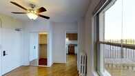 This single-floor residence offers a straightforward and functional layout with warm hardwood flooring throughout the main living area. A ceiling fan with a light fixture anchors the main room, providing both ventilation and ambient lighting. Large windows allow natural light to fill the space, with views of the surrounding brick exterior. A cast-iron radiator positioned near the window provides classic, reliable heating.

The living area connects directly to two notable spaces: a walk-in closet and a compact kitchen. The walk-in closet, accessible through a standard door, features built-in shelving and offers generous storage capacity. The kitchen is efficiently designed with warm wood cabinetry on both upper and lower sections, a stainless steel sink, a gas range, and a dishwasher. Under-cabinet lighting enhances the functional workspace.

White walls and clean crown molding throughout the unit contribute to a crisp, neutral aesthetic. The open sightlines between the main room, closet, and kitchen create a sense of connectivity across the floor plan. Overall, the space presents a practical and comfortable layout suited for everyday living, with classic architectural details that add character to its clean, modern finish.