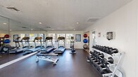 Located at 2848 Caribbean Isle Blvd in Melbourne, Florida, this fitness center offers a well-appointed exercise room designed to support a comprehensive workout routine. The space features a durable rubber-tile floor and a textured ceiling with recessed lighting, creating a clean and functional environment. Large windows along one wall flood the room with natural light and provide pleasant outdoor views, while full-length wall mirrors amplify the sense of space and allow users to monitor their form during exercise. The cardio section is equipped with multiple treadmills and an upright stationary bike, all from recognized fitness brands. A multi-station cable machine with a flat bench provides options for strength and resistance training, while a tiered dumbbell rack stocked with a full range of free weights supports a variety of exercises. A wall-mounted stability ball rack holds an assortment of medicine and exercise balls, rounding out the functional training options. Posted fitness center rules and informational signage contribute to an organized atmosphere. The room is accessed through a dedicated entryway, reinforcing its role as a standalone facility. Overall, the space is efficiently laid out to maximize usability and accommodate a diverse range of fitness activities.
