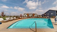 Located at 2255 E 104th Ave in Thornton, Colorado, this generously sized outdoor amenity space serves as a central gathering hub for its residential community. The centerpiece is a striking, curved resort-style swimming pool with vivid turquoise water, offering ample room for leisure and recreation. Surrounding the pool is a spacious concrete sun deck lined with rows of cushioned lounge chairs and bean bag seating, providing abundant space for relaxing under Colorado's open skies.

The pool area is enclosed by black iron fencing, ensuring both safety and an unobstructed view of the surrounding community architecture. The buildings nearby reflect a contemporary design aesthetic, combining warm wood siding, white board-and-batten panels, and stone veneer bases, accented by bold navy blue and neutral tones. Landscaping throughout features native grasses, decorative rock, ornamental trees, and shrubs that add color and texture to the environment.

A clubhouse structure anchors one side of the pool deck, featuring a prominent gabled roofline clad in warm wood tones, large windows for natural light, and a covered outdoor patio. Additional seating areas with umbrellas and lounge furniture are distributed across the deck, offering a variety of social and relaxation settings. The overall ambiance is vibrant, community-focused, and thoughtfully designed for outdoor enjoyment.