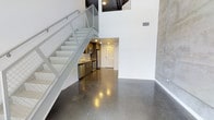 This two-story loft offers a striking industrial-modern aesthetic defined by polished concrete floors that reflect light across the open main level. The double-height ceiling creates an expansive, airy atmosphere, with crisp white walls contrasting against raw concrete accents that lend an urban character to the space. A metal-railed staircase with a mesh panel design ascends to the upper level, doubling as a visual centerpiece while tucking the kitchen neatly beneath it. The kitchen is efficiently designed with stainless steel appliances, including a refrigerator, range, and dishwasher, along with recessed lighting that keeps the area bright and functional. The open layout of the main level allows for flexible living and dining arrangements, with the polished floors adding a sleek, cohesive finish. The upper floor, accessible via the staircase, provides a sense of separation ideal for a bedroom retreat. Recessed lighting throughout ensures consistent illumination, while the architectural details — from exposed concrete walls to the industrial stair design — give this one-bedroom, one-bathroom space a distinctly contemporary character.