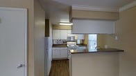 This single-story space centers around a practical kitchen designed for everyday functionality. The kitchen features white cabinetry that lines the walls, providing ample storage, paired with dark countertops that offer a subtle contrast. A white refrigerator and electric range are included, seamlessly integrated into the layout. A pass-through bar counter connects the kitchen to the adjoining living area, creating an open flow between spaces and offering additional surface area for dining or food preparation. The flooring throughout is a warm-toned hardwood laminate, adding a grounded, cohesive look. Crown molding along the upper trim adds a touch of detail to the otherwise straightforward design. A mirrored backsplash runs along the wall, visually expanding the kitchen space. Natural light filters in through a window near the range, brightening the cooking area. The textured ceiling and neutral wall tones contribute to a modest, understated ambiance throughout the space.