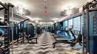 Located at 1909 Walnut St in Philadelphia's vibrant Rittenhouse Square neighborhood, this contemporary fitness center is a well-appointed, single-floor facility designed with both functionality and aesthetic appeal in mind. The space is immediately distinguished by its bold chevron-patterned carpet flooring in shades of gray and white, which runs throughout and lends the interior a dynamic, modern character.

The main workout area is expansive and generously equipped, featuring a full row of Echelon treadmills and elliptical machines positioned alongside large floor-to-ceiling windows that flood the space with natural light and offer sweeping views of the Philadelphia skyline. A dedicated strength training zone houses a comprehensive selection of cable machines, weight benches, and a full dumbbell rack, all arranged for efficient use of the floor space.

A separate studio area, enclosed by sleek glass-panel sliding doors, provides space for cycling bikes, stair climbers, and functional training with equipment such as stability balls. The entryway features a sophisticated design with textured blue-gray wall coverings, a geometric honeycomb shelving unit, and a distinctive starburst ceiling fixture, setting an elevated tone upon arrival. Cove lighting along the ceiling perimeter and recessed fixtures throughout create a well-lit, polished ambiance across the entire facility.