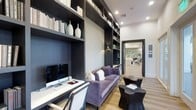 This refined single-floor space exudes a contemporary, upscale aesthetic defined by thoughtful design and curated details. The standout feature is a dramatic library lounge area where floor-to-ceiling built-in shelving in a deep charcoal finish lines the walls, housing an artful arrangement of books, decorative vases, and accent pieces. The shelving is complemented by a textured accent wall panel that adds depth and visual interest. A sleek white workstation desk is positioned along one side, complete with a desktop computer and tasteful accessories, making the space as functional as it is stylish. A plush tufted sofa in a rich lavender velvet anchors the seating area, accented by coordinating throw pillows and paired with sculptural, organically-shaped side tables in a dark finish. Recessed lighting and wall-mounted sconces provide warm, layered illumination throughout. Light wood flooring runs underfoot, offering a warm contrast to the bold dark shelving and trim. A wide doorway leads into an adjacent area, visible in the background, hinting at additional well-appointed rooms beyond. The overall ambiance is sophisticated and inviting, blending modern design sensibility with comfortable, livable elegance.