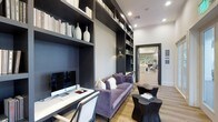 This refined single-floor space exudes a contemporary, upscale aesthetic defined by thoughtful design and curated details. The standout feature is a dramatic library lounge area where floor-to-ceiling built-in shelving in a deep charcoal finish lines the walls, housing an artful arrangement of books, decorative vases, and accent pieces. The shelving is complemented by a textured accent wall panel that adds depth and visual interest. A sleek white workstation desk is positioned along one side, complete with a desktop computer and tasteful accessories, making the space as functional as it is stylish. A plush tufted sofa in a rich lavender velvet anchors the seating area, accented by coordinating throw pillows and paired with sculptural, organically-shaped side tables in a dark finish. Recessed lighting and wall-mounted sconces provide warm, layered illumination throughout. Light wood flooring runs underfoot, offering a warm contrast to the bold dark shelving and trim. A wide doorway leads into an adjacent area, visible in the background, hinting at additional well-appointed rooms beyond. The overall ambiance is sophisticated and inviting, blending modern design sensibility with comfortable, livable elegance.