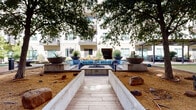 This expansive courtyard amenity space is set within a contemporary multi-story residential complex, offering a thoughtfully designed outdoor environment for recreation and relaxation. The grounds are anchored by a manicured putting green and an adjacent bocce ball court with a decomposed granite surface, providing ample space for leisure activities. String lights are strung overhead throughout the courtyard, lending a warm ambiance during evening hours.

A central fire pit lounge features a built-in concrete seating area outfitted with blue cushioned sofas and accent pillows, framed by large decorative planters and mature shade trees. A separate covered outdoor living pavilion with a wood-paneled accent wall houses a mounted flat-screen television and lounge seating, creating an inviting space for gathering.

Wide, light-tiled walkways wind through the courtyard, bordered by raised concrete planters filled with ornamental grasses, flowering shrubs, and seasonal ground cover. Open lawn areas provide additional green space, while mature trees offer natural shade along the perimeter paths. The building's contemporary white facade, balconied units, and large windows overlook the courtyard, creating a seamlessly integrated indoor-outdoor living environment filled with natural light and greenery.