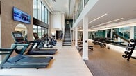This modern, two-story fitness center offers a comprehensive workout environment spread across two generously sized floors. The main level is the largest of the two, featuring soaring double-height ceilings and expansive floor-to-ceiling windows that flood the space with natural light and provide open views of the surrounding greenery and courtyard. This level houses a well-equipped cardio zone lined with treadmills, stationary bikes, rowing machines, and elliptical trainers, many outfitted with individual screens. A dedicated strength training area features a full rack of dumbbells and cable machines, while accent walls in concrete and teal tones add a contemporary aesthetic against the light hardwood flooring. A glass-railed staircase connects to the upper level, which offers additional workout areas and an overlooking mezzanine view of the main floor. A separate, enclosed functional training studio is equipped with a ceiling-mounted rig, TRX suspension trainers, a heavy punching bag, kettlebells, medicine balls, and resistance equipment, all set against mirrored walls. Throughout the facility, the clean white architectural palette, recessed lighting, and open layout create a bright, motivating atmosphere suited for a full range of fitness activities.