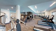 Located at 5080 Camino Del Arroyo in San Diego, this two-story fitness center delivers a comprehensive and well-equipped workout environment designed to accommodate a wide range of training styles. The facility features a modern, clean aesthetic throughout, with soft teal-accented ceilings, neutral carpeted flooring in primary workout areas, and linear LED lighting that keeps each zone bright and functional.

The main floor houses an expansive cardio zone lined with rows of TRUE-brand treadmills, ellipticals, and stair climbers, complemented by floor-to-ceiling mirrors that enhance the sense of space. Large-screen televisions are mounted along the walls for entertainment during workouts. A dedicated free weights area includes a full dumbbell rack, adjustable benches, cable machines, and additional strength equipment. A teal accent wall and built-in shelving with a mini-refrigerator provide a welcoming amenity nook near the staircase. Stair climber machines and functional training equipment round out the ground level.

A striking metal staircase with horizontal railing leads to the upper level, which features a more intimate layout. This floor includes a red cable-and-functional-training rig, TRX suspension systems, rowing machines, and open floor space ideal for stretching or dynamic movement. Two dedicated boxing rooms—one with a heavy bag and one with a speed bag and interactive striking unit—are finished with hardwood-style flooring. Large windows on the upper level offer outdoor views, filling the space with natural light.