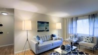 This single-floor space offers a clean, well-appointed interior designed with a cohesive nautical aesthetic. The living area serves as the heart of the home, featuring warm hardwood flooring that anchors the room with a sense of warmth and comfort. Neutral, softly painted walls provide a versatile backdrop that enhances the light-filled ambiance created by large windows dressed in flowing blue-toned curtains.

The living room is thoughtfully arranged around a plush, tufted light grey sofa accented with navy and patterned throw pillows, conveying a relaxed yet refined atmosphere. Two sleek tripod floor lamps flank the seating area, providing both functional and ambient lighting. A glass-topped coffee table reflects light and contributes to the open, airy feel of the space.

A standout decorative element is a bold nautical painting depicting a sailboat at sea, hung prominently on the main wall, reinforcing the coastal theme. An armchair and additional accent pieces near the window complete the seating arrangement, making the space ideal for relaxing or entertaining.

Overall, this space strikes a balance between casual comfort and tasteful style, with its maritime-inspired décor, natural light, and warm wood flooring creating an inviting and cohesive environment throughout.