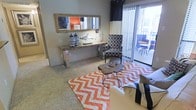 This single-floor space showcases a thoughtfully designed living area that blends contemporary style with bold, eye-catching décor. The living room serves as the focal point of the space, featuring a striking orange and white chevron area rug that anchors the room with a burst of color and energy. A sleek console table adorned with decorative vases and sculptural accents lines one wall beneath a large, ornate framed mirror, adding depth and visual interest to the space. A patterned accent chair in a classic geometric print complements the eclectic design aesthetic, while a rustic live-edge coffee table introduces a natural, organic contrast to the modern surroundings. Neutral-toned carpeting and soft grey walls provide a balanced backdrop that allows the vibrant furnishings to take center stage. Large sliding glass doors flood the living area with natural light and open directly onto a private outdoor balcony, where additional seating creates an inviting extension of the interior living space. The seamless indoor-outdoor connection enhances the overall sense of openness and airiness throughout the home. The cohesive design and carefully curated details make this a warm, stylish, and functional living environment.