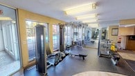 This fitness and wellness facility is thoughtfully designed to provide a comprehensive workout and recovery experience within a single-floor layout. The main exercise area is outfitted with professional-grade strength training equipment, including a multi-station cable machine and various resistance apparatus from Life Fitness, arranged across rubber flooring that provides both safety and durability. Large leaded-glass windows line the walls, flooding the space with natural light and offering views of a pool area and surrounding greenery, creating an inviting and energizing atmosphere. Full-length mirrors along one wall amplify the sense of space and allow users to monitor their form. Overhead fluorescent lighting supplements the natural light, ensuring the area remains well-lit at all times. The textured ceiling and warm yellow accent walls contribute to a comfortable, functional ambiance. Adjacent to the gym floor, a dedicated sauna room is clearly marked and accessed through a solid wood door, offering a private space for heat therapy and relaxation. The facility also includes a shower area equipped with a curtained stall and overhead wire shelving for storage, providing a convenient post-workout rinse. Louvered ventilation panels are integrated into the design to maintain proper airflow throughout the space.