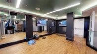 This fitness space is designed with a contemporary aesthetic, offering a well-appointed environment for exercise and training. The room features warm hardwood flooring throughout, providing a clean and functional surface suited for a variety of workout activities. Bold dark structural columns are positioned throughout the space, adding an architectural contrast against the lighter flooring and mirrored walls.

Full-length mirrors line one wall, visually expanding the room and allowing users to monitor their form during exercise. Multiple large flat-screen televisions are mounted at eye level, ensuring entertainment or news access from various points in the room. Recessed LED strip lighting runs along the dropped ceiling perimeter, emitting a soft ambient glow that complements the modern design, while in-ceiling speakers suggest an integrated audio system.

Fitness accessories are present throughout, including stability balls, a BOSU balance trainer, and additional small equipment, indicating the space supports functional and balance-focused training. A secondary area visible through the mirrors suggests additional equipment or storage nearby. The overall ambiance is sleek, modern, and well-lit, creating a focused and professional workout environment.