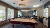 This single-floor amenity space functions as a combined game room and social lounge, designed with a cohesive, contemporary aesthetic. The centerpiece is a full-size billiard table with rich mahogany legs and a tan felt surface, positioned beneath an elegant brass chain-hung pendant light that serves as a focal point for the room. Patterned carpet tiles in muted earth tones cover the flooring throughout, adding texture while complementing the warm taupe and gray wall palette.

Large windows along multiple walls invite generous natural light and offer views of an outdoor courtyard featuring arched architectural details. A full-length mirror on one wall visually expands the space, reflecting both the game area and the adjacent lounge zone. The lounge area features a comfortable upholstered sofa accented with decorative pillows, flanked by wall sconces and a bold abstract artwork, creating an intimate retreat within the broader room.

Multiple high-top bistro tables with wrought-iron chairs are arranged throughout, providing flexible seating options for socializing. A wall-mounted flat-screen television is positioned for easy viewing from various points in the room, and a dedicated coffee and refreshment station offers added convenience. A cue rack and additional storage complete the practical amenities of this thoughtfully designed communal space.
