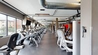 Located at 6250 Griffin Rd in Davie, FL, this fitness facility spans a generously sized single floor, delivering a fully equipped gym environment designed to support a wide range of workout routines. The space features an open, industrial aesthetic defined by exposed ductwork and conduit running along the ceiling, complemented by a neutral gray color palette punctuated by bold accent walls in vivid orange-red, adding energy to the environment.

The cardio zone is a standout feature, housing an extensive row of treadmills, ellipticals, stair climbers, and stationary bikes, all positioned along window-lined walls that invite natural light and provide outdoor views of the surrounding Davie area. Wall-mounted flat-screen televisions are stationed throughout, offering entertainment options during workouts.

A dedicated strength training section includes multiple cable machines, power racks, benches, and free weights arranged on durable rubber flooring, with full-length mirrors amplifying the sense of space. A punching bag station adds a functional training dimension. Smooth concrete-style flooring runs through the cardio areas, ensuring easy navigation between equipment zones.

Structural columns are integrated throughout the layout, contributing to the architectural character while defining distinct workout areas. The overall ambiance is modern, functional, and professional, reflecting a well-organized facility built for high-capacity use.