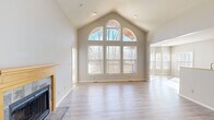 This single-floor home is defined by its dramatic architectural character, most notably in the expansive living room where soaring vaulted ceilings rise to a peak framed by a striking arched window flanked by tall double-pane windows. This feature wall floods the space with natural light and offers views of surrounding mature trees, creating an airy, open atmosphere. Light wood-look flooring runs throughout the living area, complementing neutral gray-toned walls. A tile-surround fireplace with a natural wood mantel anchors one side of the room, adding warmth and a focal point to the space. The kitchen is well-appointed with honey-toned wood cabinetry, granite-look countertops, tile backsplash, and black appliances including a range, microwave, and refrigerator, all set on light hardwood-style flooring. The bedrooms feature soft carpeting; one includes a bay window configuration that brings in abundant natural light, while another showcases vaulted ceilings. An upper loft-style area with a skylight and wood railing detail adds a bonus flexible space. The full bathroom offers a single-sink vanity with wood cabinetry and a tub-shower combo. A dedicated laundry room with a full-size washer and dryer completes this thoughtfully designed home.