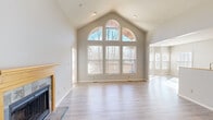 This single-floor home is defined by its dramatic architectural character, most notably in the expansive living room where soaring vaulted ceilings rise to a peak framed by a striking arched window flanked by tall double-pane windows. This feature wall floods the space with natural light and offers views of surrounding mature trees, creating an airy, open atmosphere. Light wood-look flooring runs throughout the living area, complementing neutral gray-toned walls. A tile-surround fireplace with a natural wood mantel anchors one side of the room, adding warmth and a focal point to the space. The kitchen is well-appointed with honey-toned wood cabinetry, granite-look countertops, tile backsplash, and black appliances including a range, microwave, and refrigerator, all set on light hardwood-style flooring. The bedrooms feature soft carpeting; one includes a bay window configuration that brings in abundant natural light, while another showcases vaulted ceilings. An upper loft-style area with a skylight and wood railing detail adds a bonus flexible space. The full bathroom offers a single-sink vanity with wood cabinetry and a tub-shower combo. A dedicated laundry room with a full-size washer and dryer completes this thoughtfully designed home.