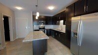 This contemporary single-floor space is anchored by a thoughtfully designed kitchen that serves as the heart of the unit. Rich, dark espresso cabinetry lines the walls from base to upper level, providing generous storage while creating a bold, sophisticated aesthetic. The countertops throughout feature light-toned granite with subtle veining, offering a striking contrast against the dark cabinetry. A central kitchen island extends the prep and entertaining space, topped with the same granite surface and illuminated by stylish pendant lights that hang from above, adding warmth and focused lighting. The kitchen is fully equipped with stainless steel appliances, including a built-in microwave, electric range, and a French door refrigerator with an ice and water dispenser. A stainless steel undermount sink is positioned along the perimeter counters, accompanied by a modern faucet. A classic white subway tile backsplash runs along the walls, adding texture and brightness to balance the darker tones. The flooring throughout is a light-toned wood-look material, enhancing the open and airy feel of the space. Clean white walls and smooth ceilings complete the modern, move-in-ready aesthetic of this well-appointed unit.
