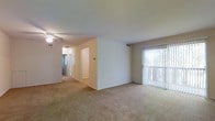 This single-floor residence offers a clean, functional layout designed for comfortable everyday living. The main living area is generously proportioned, featuring wall-to-wall carpeting in a neutral beige tone that adds warmth and softness underfoot. A large sliding glass door fitted with vertical blinds allows abundant natural light to fill the space while offering a view of the greenery outside, creating a bright and airy atmosphere.

The room features smooth white walls and a ceiling-mounted light fixture, contributing to a simple, understated aesthetic. A centrally positioned partial wall divides the living space, providing a subtle sense of structure without compromising the open feel of the layout.

A hallway visible from the main living area leads to the additional rooms of the unit, suggesting a thoughtful floor plan that separates the living and private spaces. The corridor is well-lit and carpeted, maintaining a consistent design language throughout. Ceiling vents are present throughout, indicating a central HVAC system for year-round climate control. Overall, the space presents a practical and move-in-ready environment with a neutral palette that offers versatility for a variety of interior styles.