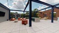 This contemporary building offers a well-appointed interior and a standout rooftop terrace that serves as a central amenity. The interior hallways are thoughtfully designed with a two-tone wall treatment — light grey above and deep charcoal wainscoting below — complemented by patterned carpet flooring in blue and white geometric tones. Recessed lighting illuminates the length of the corridor, while large windows along the hallway bring in natural light and offer views of the adjacent brick building exterior. Glass-paned doors with push-bar hardware provide access between interior spaces, maintaining an open and connected feel throughout.

The rooftop terrace is a highlight of the property, featuring large-format porcelain tile flooring and a bold navy steel pergola structure strung with bistro lighting, creating an inviting atmosphere for both day and evening use. The outdoor space is furnished with plush lounge seating in warm terracotta tones, complemented by teal bar stools arranged along a concrete bar counter. Additional seating options, including wicker accent chairs, are positioned throughout. Surrounding views of mature trees, neighboring rooftops, and open sky enhance the open-air ambiance. Corrugated metal utility enclosures are discretely integrated into the terrace layout, maintaining a clean and organized aesthetic overall.