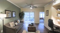 This single-floor residence in Dallas, Texas, offers a thoughtfully designed living space with a welcoming and comfortable ambiance. The living room serves as the heart of the home, showcasing rich dark hardwood flooring that contrasts beautifully with the soft, neutral wall tones accented by a bold sage green feature wall. Crown molding adds a refined architectural touch throughout, while a ceiling fan provides both style and practical comfort. Large sliding glass doors flood the space with natural light and open onto an outdoor balcony, seamlessly connecting the interior to the outdoors. The living area provides ample room for relaxed seating arrangements and is complemented by tasteful décor, including framed artwork and lush indoor greenery. A glimpse into the adjacent bedroom reveals plush carpeting and continued access to natural light, suggesting a cohesive and comfortable design throughout the home. The overall layout flows naturally from one space to the next, offering a blend of functionality and style well-suited to everyday living in the heart of Dallas.