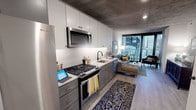 This single-floor urban residence showcases a contemporary industrial-chic design, defined by signature exposed concrete ceilings that run throughout the entire space. Light wood-tone flooring provides a warm contrast to the raw ceiling texture, establishing a cohesive aesthetic from entry to balcony.

The galley-style kitchen is well-appointed with two-tone cabinetry — white upper cabinets paired with grey lower units accented by gold hardware — complemented by a herringbone-patterned tile backsplash. Stainless steel appliances include a gas range, built-in microwave, refrigerator, and dishwasher, all set beneath under-cabinet lighting.

The open-plan living area flows naturally from the kitchen toward floor-to-ceiling glass sliding doors, which open onto a private balcony with sweeping city skyline views. The living space offers ample room for seating and entertaining while benefiting from abundant natural light.

The bedroom continues the exposed concrete ceiling motif and features expansive corner windows framing dramatic urban views, creating a distinctive and light-filled sleeping environment.

The bathroom is finished with grey shaker-style cabinetry, gold and matte black hardware, backlit mirrors, white subway tile surrounding the combination tub and shower, and large-format floor tiles, delivering a polished, modern aesthetic.