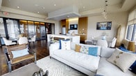 This thoughtfully designed single-floor space showcases a seamless open-concept layout that integrates living, dining, and kitchen areas into one cohesive environment. Rich dark hardwood flooring runs throughout, providing a warm, grounding contrast to the light-toned walls and recessed ceiling lighting above.

The living area features a contemporary linear fireplace set into a clean, minimalist surround, flanked by generous seating and framed by expansive floor-to-ceiling windows and glass doors. These openings flood the interior with abundant natural light while offering elevated views of the surrounding cityscape and beyond.

The dining area centers around a striking round glass-top table with a sculptural wood base, surrounded by elegant cream-upholstered chairs. Adjacent to the living space, a modern kitchen presents a white waterfall-edge island, warm wood cabinetry with integrated lighting, and stainless steel appliances — combining functionality with refined aesthetics.

Throughout, the design maintains a consistent contemporary sensibility, balancing neutral tones with carefully chosen accents, layered textiles, and curated decor. The result is a bright, polished, and welcoming space well-suited for both everyday living and entertaining.