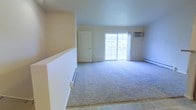 This single-floor space features an open living area finished with neutral-toned carpet that extends across the main floor, transitioning to tile flooring at the entryway. The room is kept bright and airy by a large sliding glass door that draws in natural light and provides views of the surrounding exterior. A wall-mounted air conditioning unit is positioned above the sliding door, complementing the baseboard heating system that runs along the perimeter walls for year-round climate control. The walls are painted in a clean, light neutral tone, contributing to the overall sense of openness. A standard interior door leads to an additional area or closet space, adding functional versatility to the layout. The combination of the wide carpeted area and the tile entryway provides a practical flow between zones. The space presents a straightforward, well-lit interior well-suited for comfortable everyday living.