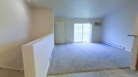 This single-floor space features an open living area finished with neutral-toned carpet that extends across the main floor, transitioning to tile flooring at the entryway. The room is kept bright and airy by a large sliding glass door that draws in natural light and provides views of the surrounding exterior. A wall-mounted air conditioning unit is positioned above the sliding door, complementing the baseboard heating system that runs along the perimeter walls for year-round climate control. The walls are painted in a clean, light neutral tone, contributing to the overall sense of openness. A standard interior door leads to an additional area or closet space, adding functional versatility to the layout. The combination of the wide carpeted area and the tile entryway provides a practical flow between zones. The space presents a straightforward, well-lit interior well-suited for comfortable everyday living.