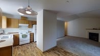 This single-floor residential space offers a practical and functional layout designed for comfortable everyday living. The kitchen is outfitted with warm-toned wooden cabinetry, a white electric range, and a full-size refrigerator, providing ample storage and prep space. A textured ceiling and a pendant barn-style light fixture overhead add a touch of character to the kitchen area. The flooring transitions from wood-look planks in the kitchen and entry zone to soft carpeting in the adjacent living area, creating a clear and natural delineation between the two spaces. A prominent partial wall separates the kitchen from the living room, offering a sense of definition while maintaining an open, connected feel throughout. The living area is generously proportioned, with neutral-toned walls that provide a versatile backdrop. Subtle recessed lighting in the living space contributes to a calm, evenly lit ambiance. Overall, the space presents a clean, move-in ready environment with a neutral palette well-suited to a variety of furnishing styles and personal aesthetics.
