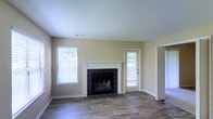 This single-story space centers around a well-appointed living area that combines comfort and classic design elements. The focal point of the room is a wood-burning fireplace framed by a crisp white mantel and surround, with dark slate-style tile extending from the firebox to the floor, creating a striking contrast against the neutral beige walls.

The flooring throughout the living area consists of hardwood-style planks in a warm, medium-toned finish, lending the space a clean and cohesive aesthetic. A textured popcorn ceiling adds a traditional touch overhead, while the white baseboards and door trim provide neat, defined lines throughout.

Natural light fills the room through multiple windows fitted with horizontal blinds, offering both brightness and privacy. A glass-paneled exterior door, positioned adjacent to the fireplace, provides access to an outdoor area and allows additional natural light to filter in. A secondary doorway on the right side of the room connects to additional areas of the space. Overall, the living area presents a functional, straightforward layout with classic residential finishes.