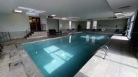 This indoor aquatic facility is designed to provide a comfortable and functional recreational environment. The centerpiece is a generously sized swimming pool with clear, teal-tinted water, framed by smooth tiled surroundings and accessible via a stainless steel entry railing. Adjacent to the main pool sits a hot tub, offering a relaxing complement to the swim area. The space is finished with neutral green-gray walls and a light-colored ceiling, contributing to a clean, open atmosphere. Natural light filters in through multiple windows along the walls, brightening the interior and offering views to the outside. The flooring throughout the pool deck is finished in a durable, slip-resistant tile, practical for a wet environment. Several lounge chairs are arranged along the perimeter, providing a comfortable space for relaxation before or after swimming. Safety signage is posted near the entrance, and a life ring is mounted on the wall for emergency use. A potted plant near the far wall adds a subtle touch of greenery. The overall ambiance is calm and utilitarian, making this facility well-suited for both leisure and light aquatic exercise.