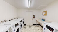 Located at 801 D St in Davis, California, this shared laundry facility offers a clean, well-organized space designed for practical, everyday use. The room is finished with a classic black-and-white diamond-patterned tile floor and bright white walls, creating a tidy and functional environment. Overhead fluorescent lighting ensures the space is well-lit throughout.

Multiple front-loading washing machines and top-loading dryers are arranged along the walls, providing ample capacity for users. The machines are modern and clearly labeled, with accessible units marked for those with mobility needs. A built-in folding counter along one wall offers a convenient surface for sorting and folding laundry, accompanied by a corkboard for posting notices and information.

A utility sink area is tucked into a corner of the room, complete with wall-mounted faucets and a trash receptacle, adding to the overall utility of the space. A fire extinguisher is mounted near the entry door, reflecting attention to safety. Ventilation is provided through a wall vent, keeping the space comfortable during use. The single-level layout and accessible design make this laundry facility a straightforward, efficiently arranged shared amenity.